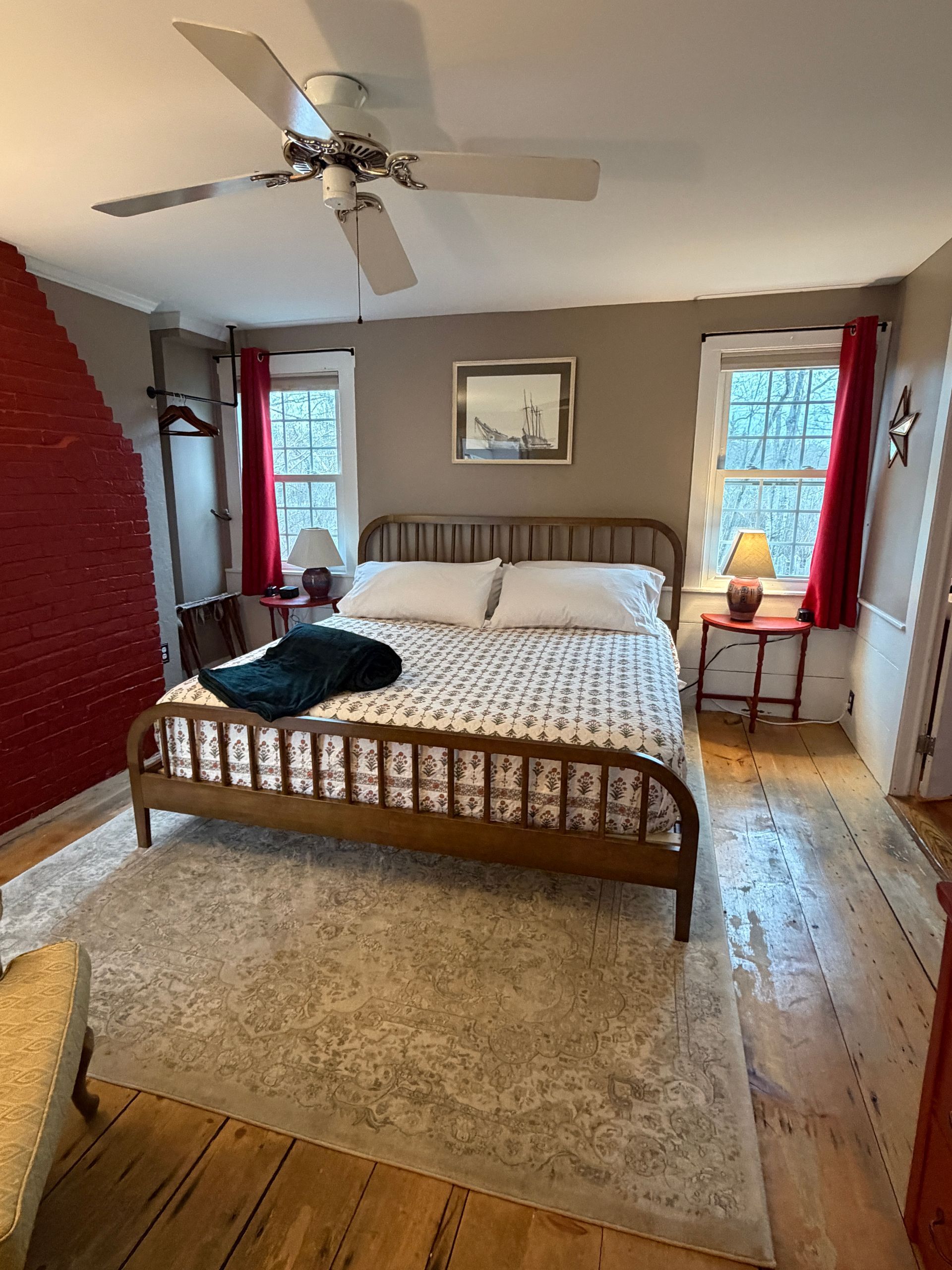 Cozy bedroom with a wooden bed, beige rug, ceiling fan, and two windows with red curtains.