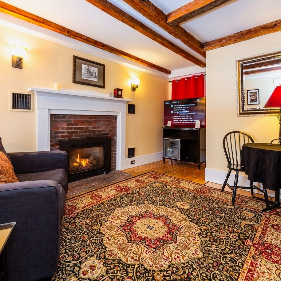 Cozy living room with fireplace, rug, furniture. Wooden beams, TV, and black-covered table with chair.