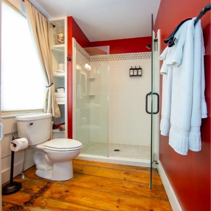 Bathroom with red walls, glass shower, wooden floor, toilet, and towels.