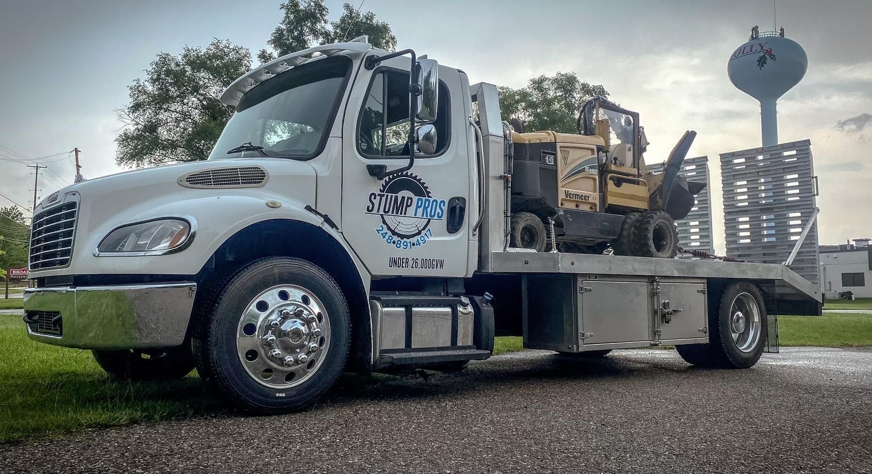 A white tow truck with a forklift on the back is parked on the side of the road.