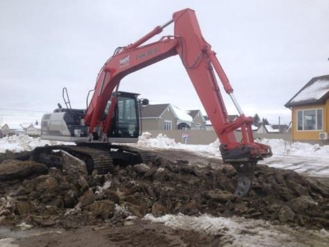 Une excavatrice rouge creuse un trou dans la terre devant une maison.