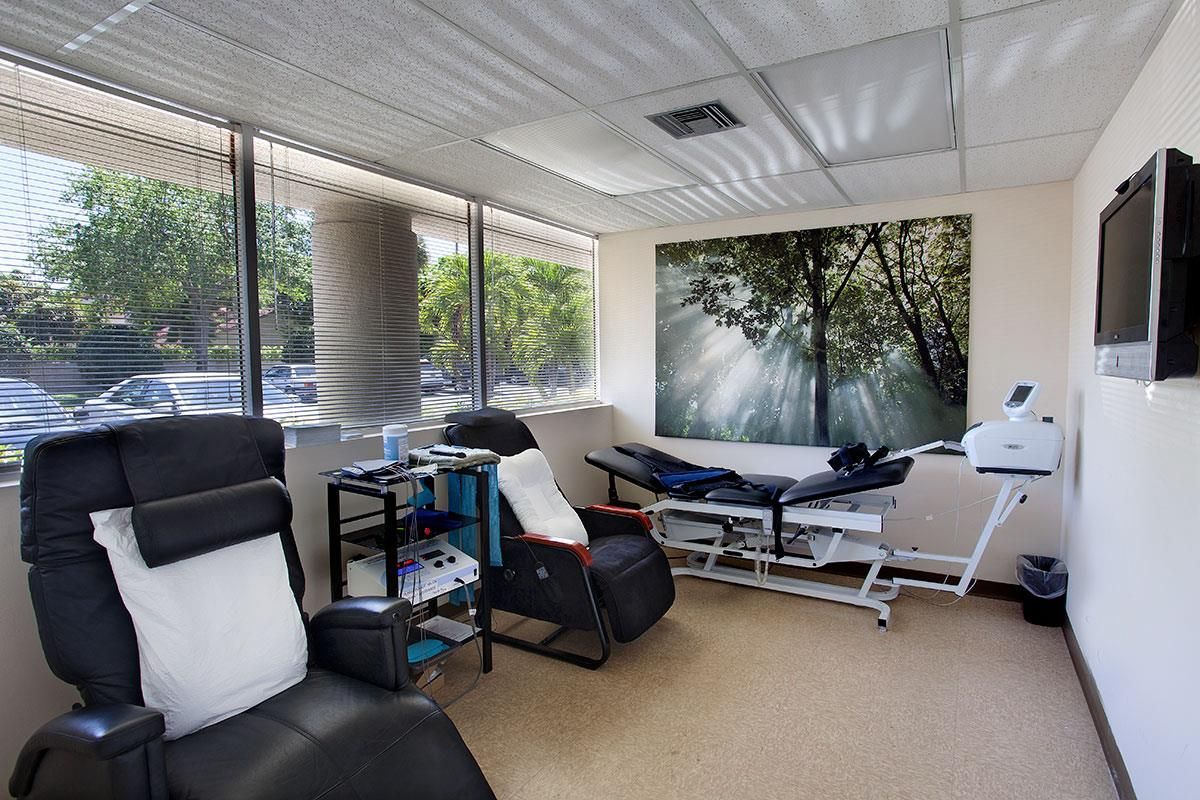 Medical room with reclining chair, treatment table, equipment, large window with trees, and nature artwork.