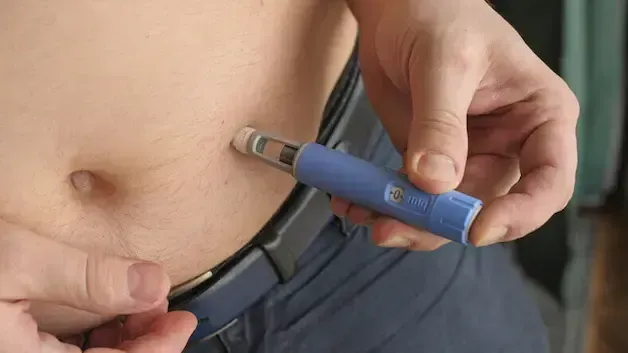 Person injecting medication into abdomen with a blue pen injector.