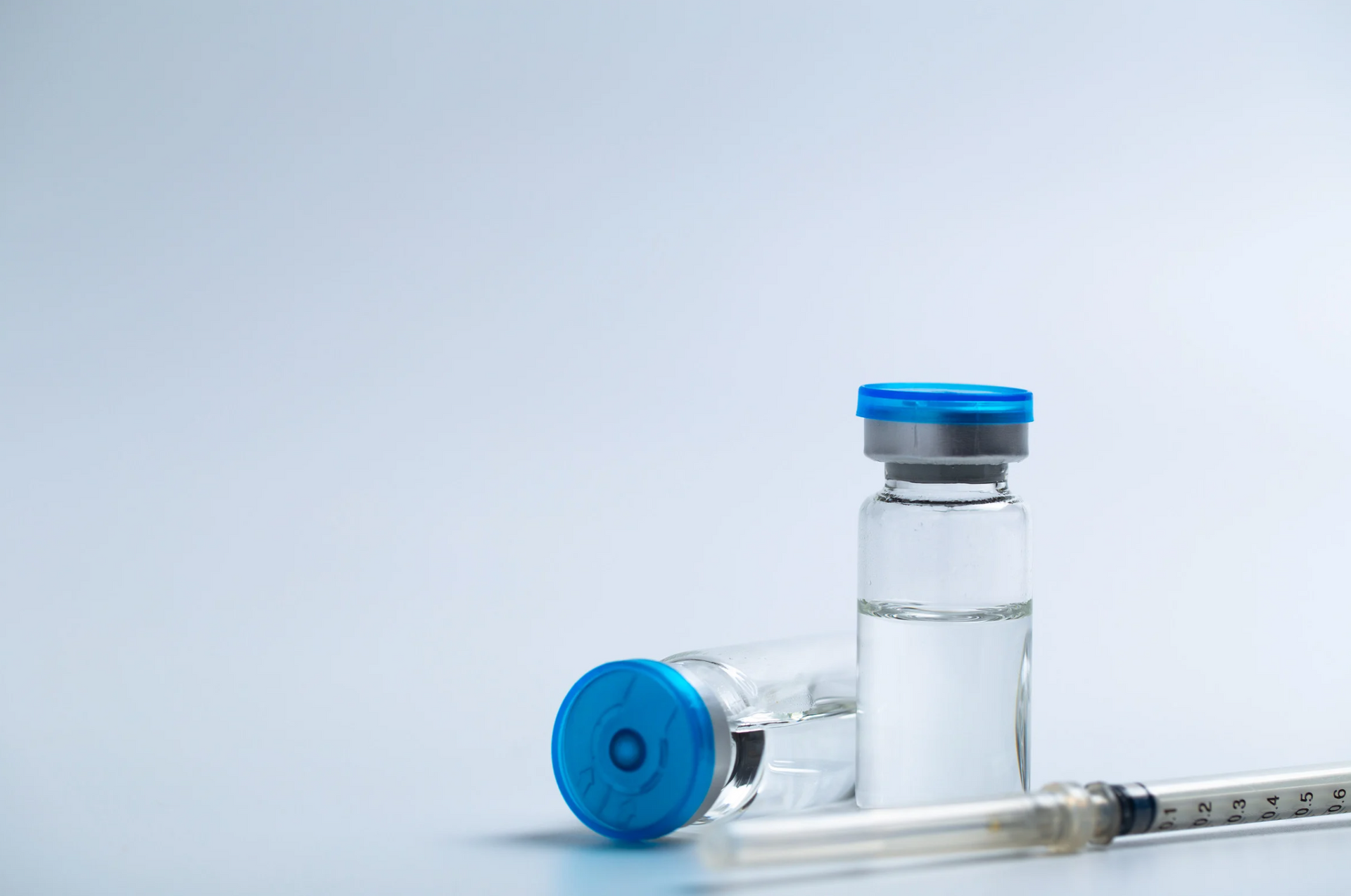 Two vaccine vials with blue caps, and a syringe on a light blue background.