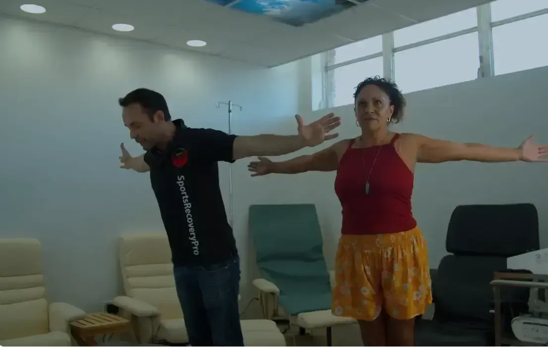Two people with arms outstretched in a room with chairs. Person on left leans forward.
