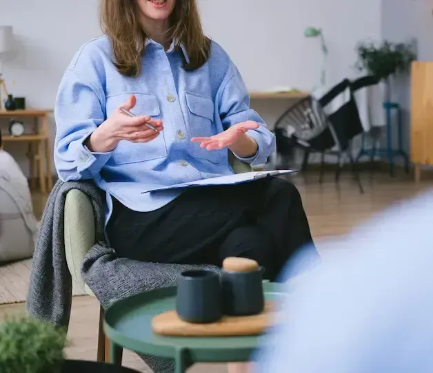 Woman in blue shirt gestures while sitting, holding a clipboard in a therapy session setting.