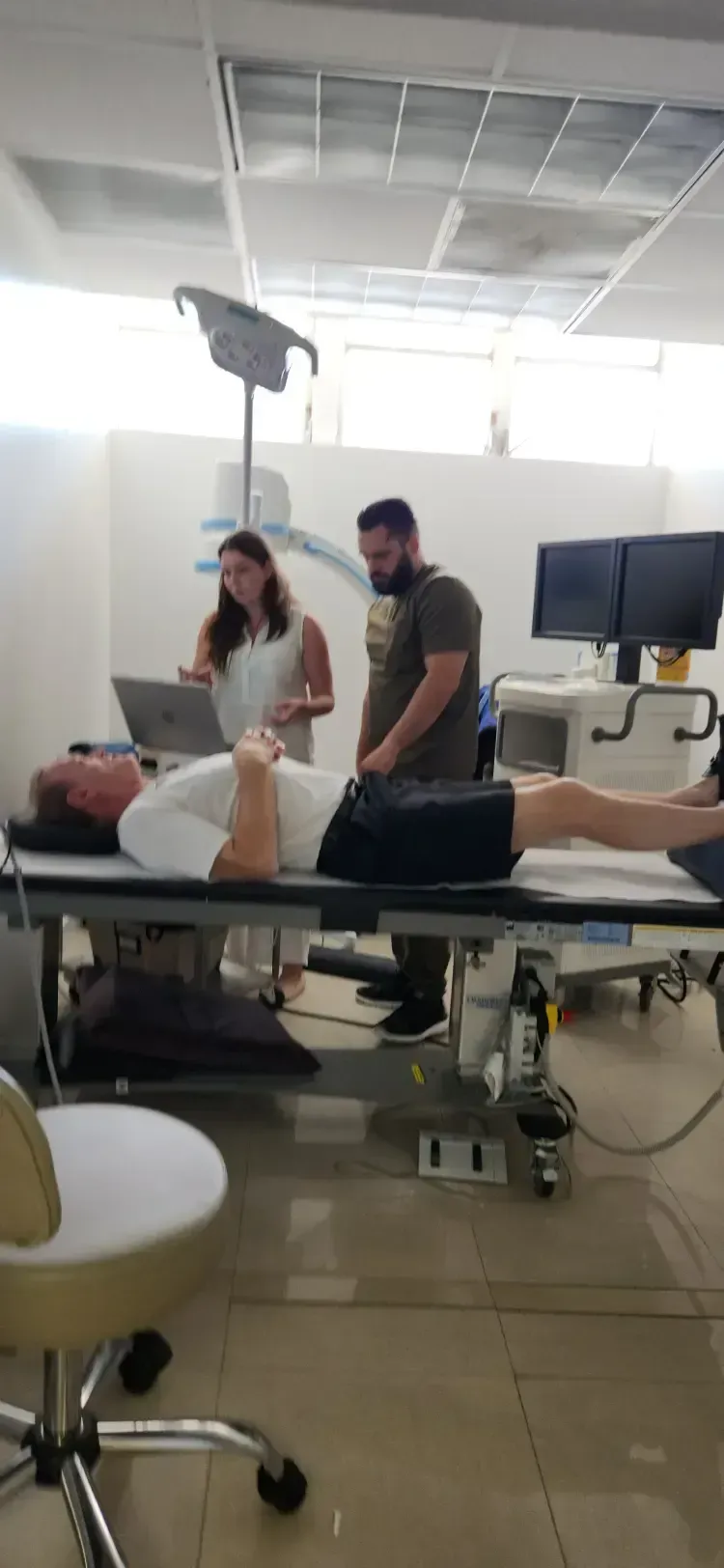 A person lying on a medical examination table with two people standing near them and a laptop.