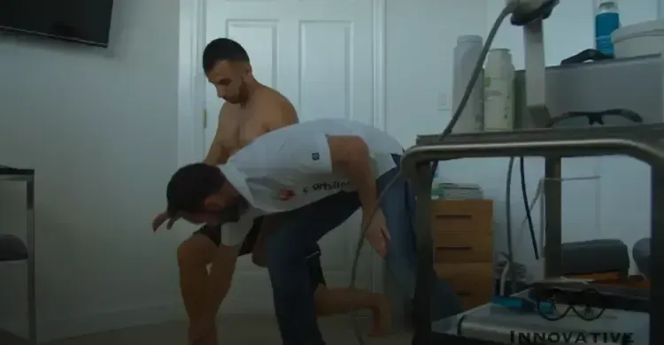 Two men in an indoor setting; one bends over while the other stands shirtless behind him, possibly providing assistance.
