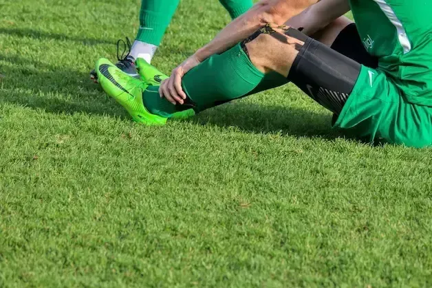 Soccer player on grass clutches leg, appearing injured, wearing green uniform.