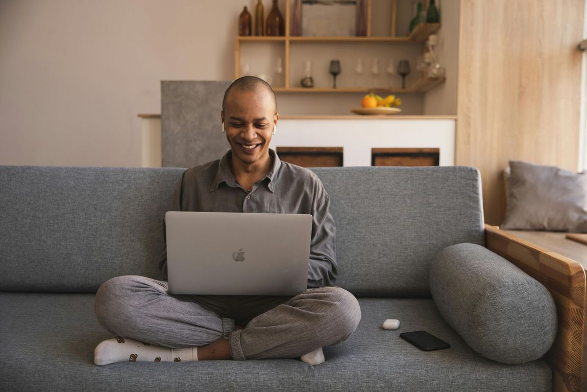 Person with shaved head smiling, working on laptop while sitting cross-legged on a gray couch in a living room.