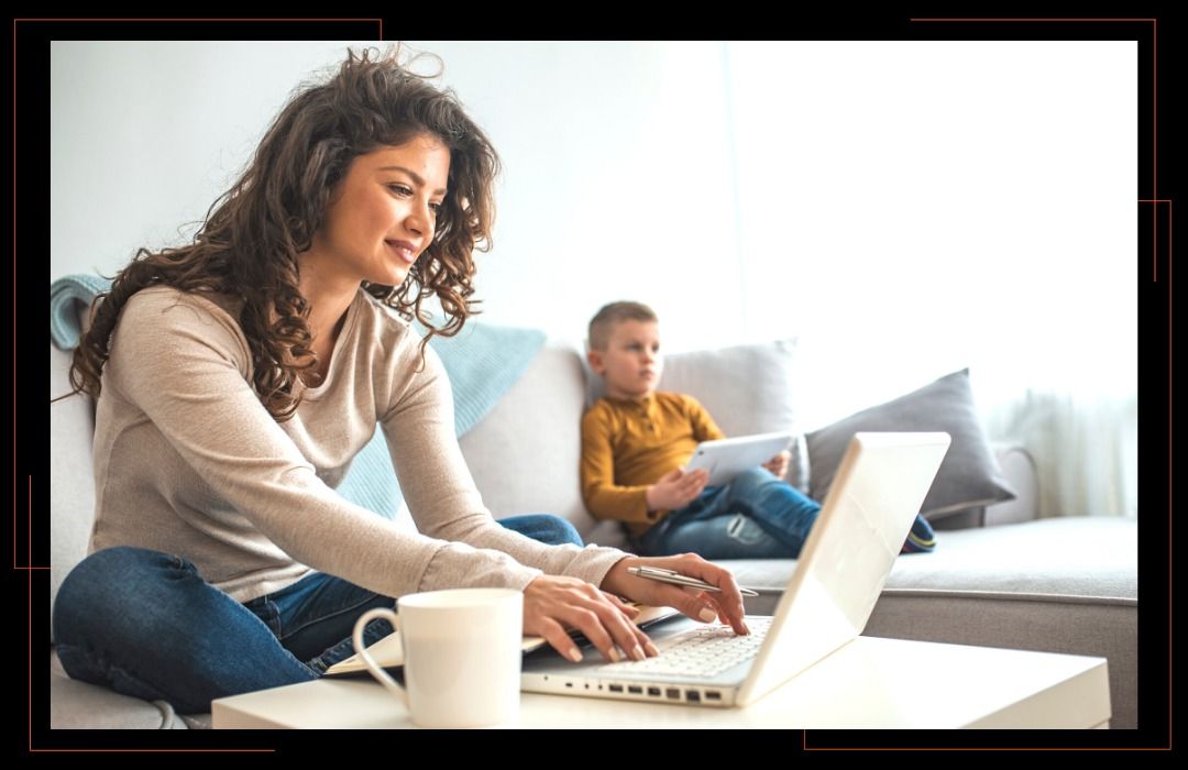 A woman is sitting on a couch using a laptop computer while a child sits on a couch.