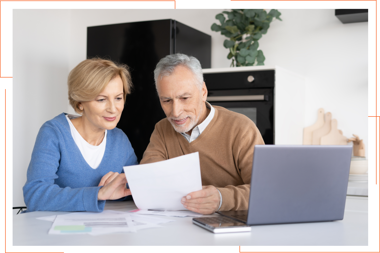 An elderly couple is sitting at a table looking at papers and a laptop.