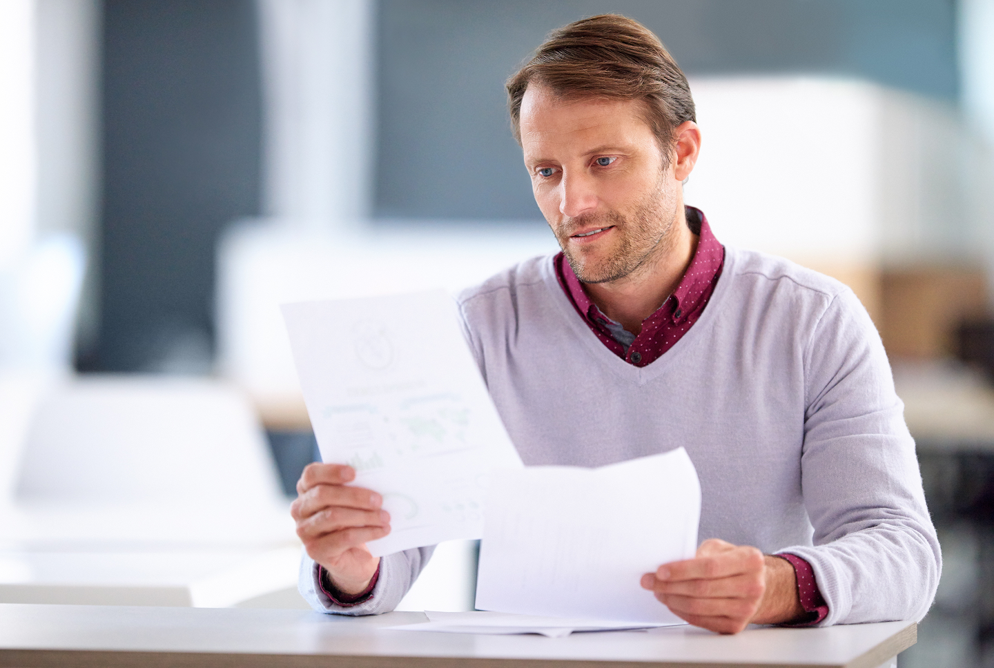 Man in a light sweater and burgundy shirt, looking at papers while sitting at a desk.