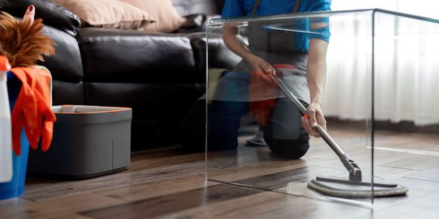 Person cleans a hardwood floor with a mop. Cleaning supplies and a couch are in the background.