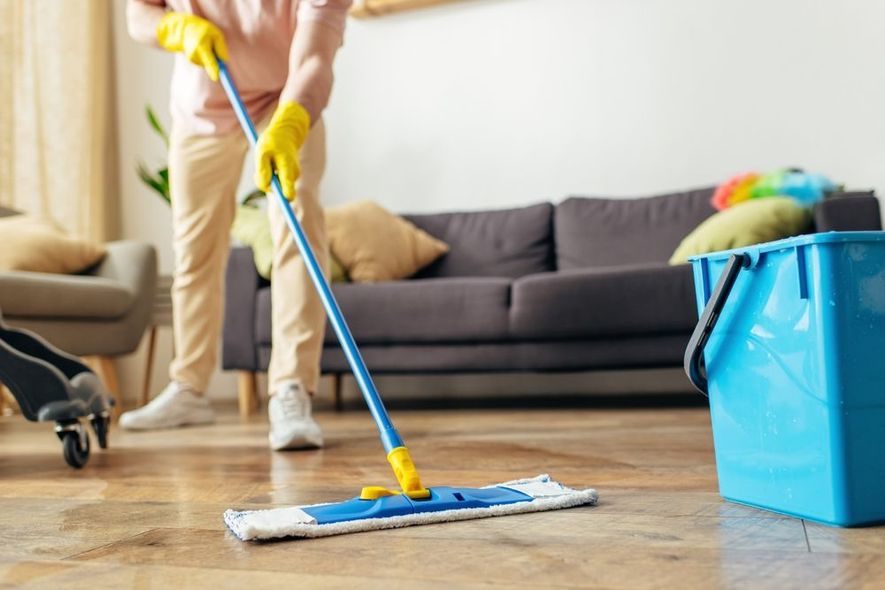 Person mopping wooden floor in living room with yellow gloves, blue bucket nearby.