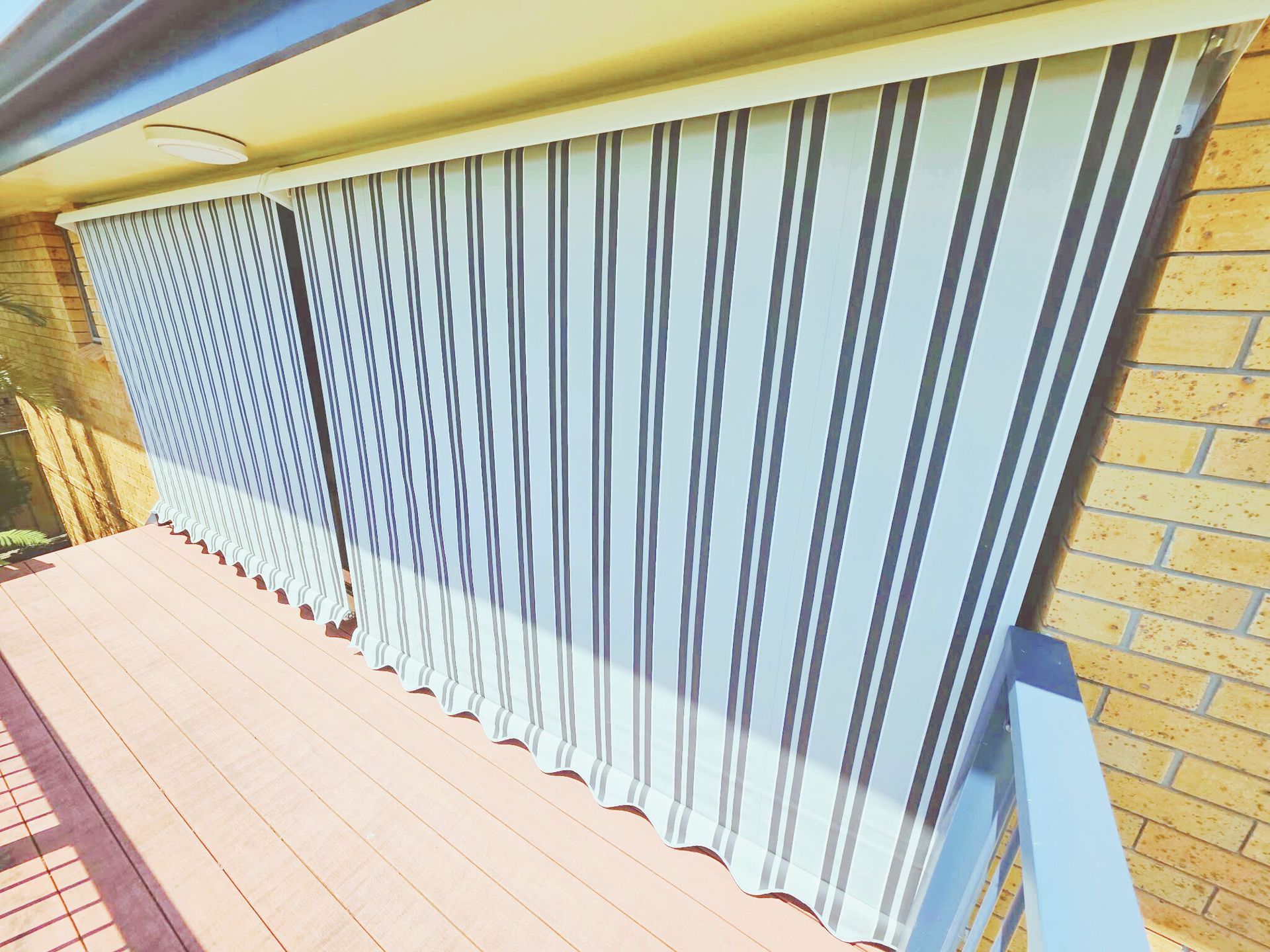 Curving walkway with overhead canopy in a park setting, blue sky, trees, and buildings in the distance.