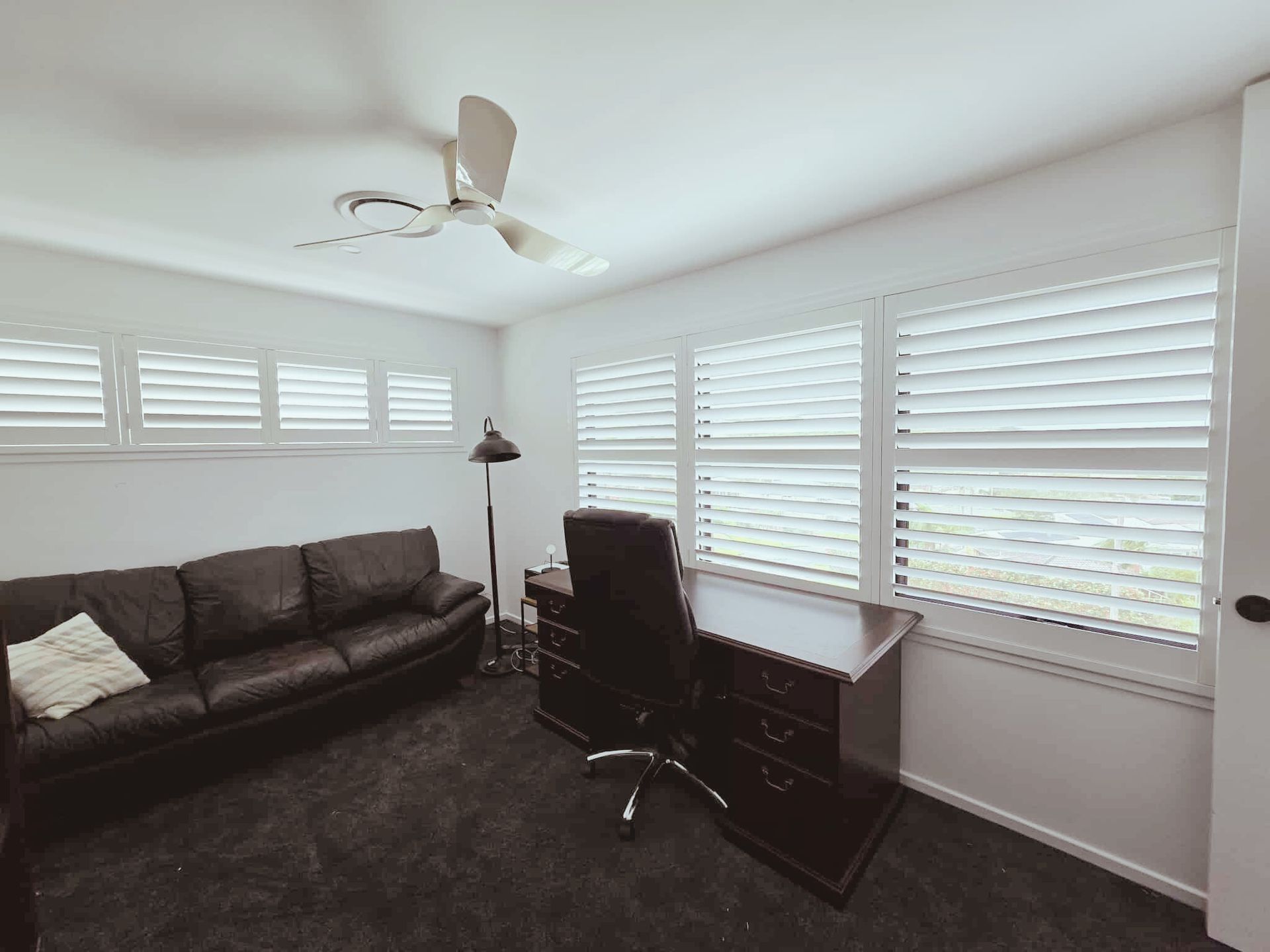 Black window blinds in a white window sill, with a pillow and vase of dried flowers.