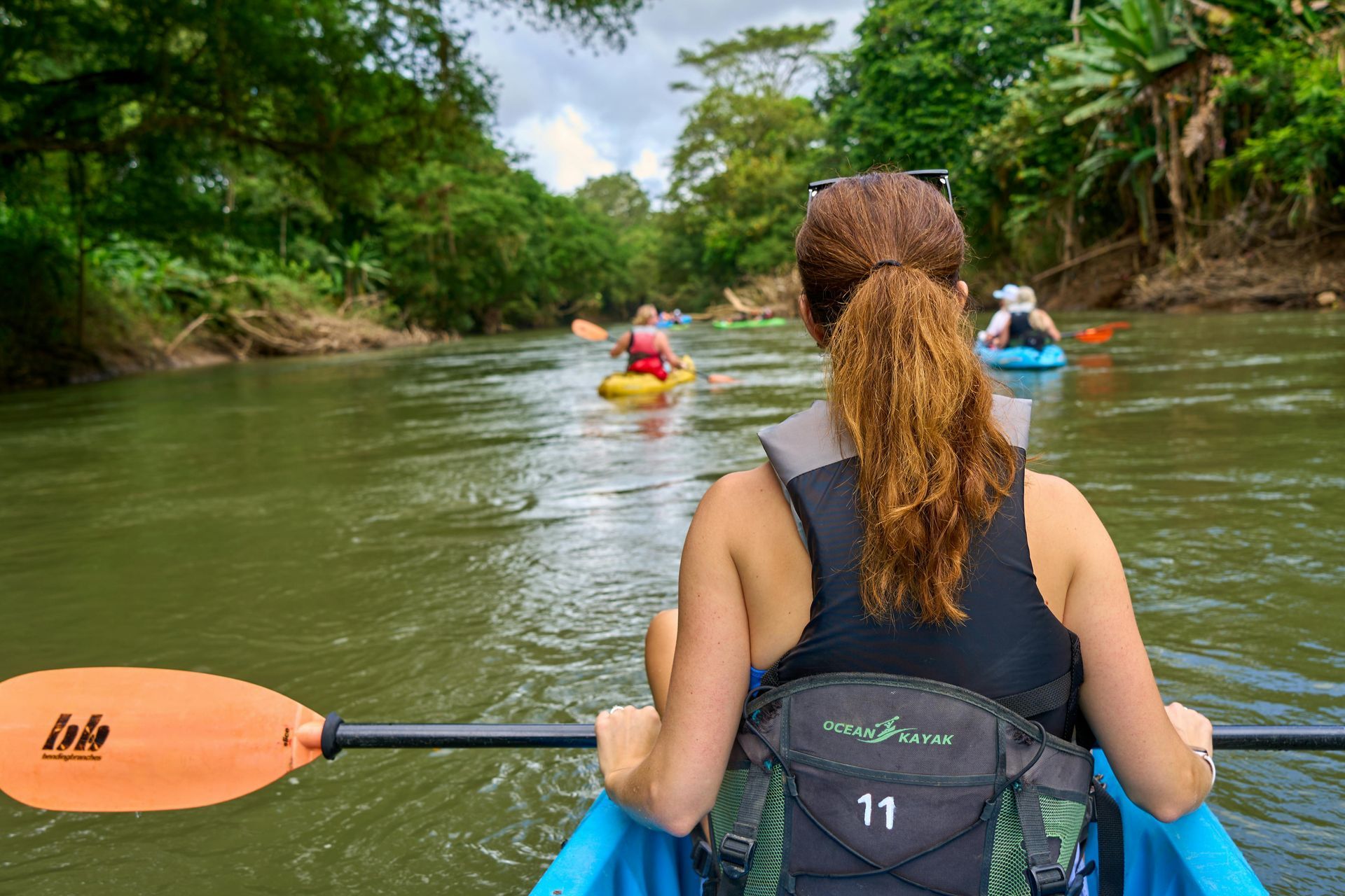 Woman kayaking on a river in Vermont, facing forward, with other kayakers ahead. Lush green trees line the riverbanks on an overcast day.