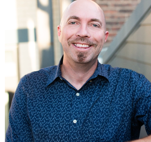 A bald man with a beard wearing a blue shirt is smiling for the camera.