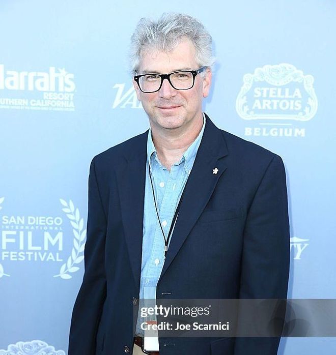 A man in a suit and glasses stands in front of a stella artois sign