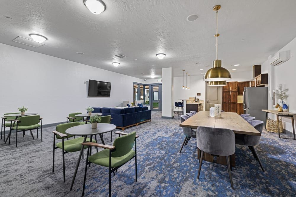A modern and spacious interior common area with mixed seating options including green chairs and round tables in the foreground, a blue velvet sofa with a matching rug in the middle, and a wooden dining table set with grey chairs under a large brass pendant light. The room has a blue patterned carpet, white walls, and a kitchenette with wood cabinetry in the background. A flat-screen television hangs on the left wall, and the space is lit with a combination of ceiling downlights and stylish hanging light fixtures at Copperline at Point Ruston in Tacoma, WA.