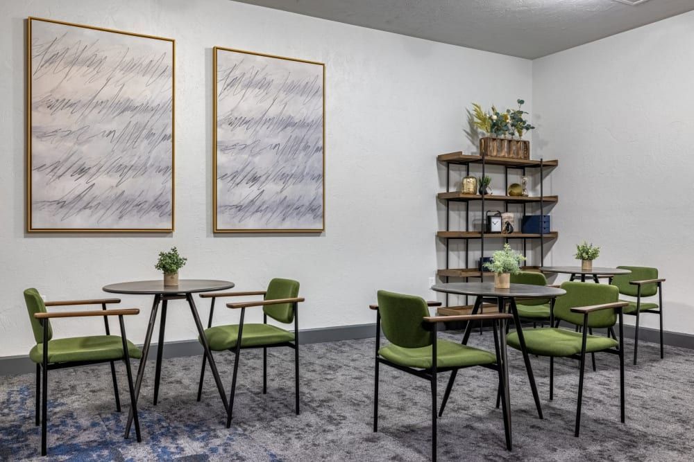 A modern waiting area with two abstract paintings framed in gold above a round wooden table flanked by green upholstered chairs with wooden arms. Across the room stands a stylish shelving unit adorned with decorative books, plants, and golden accents, creating an inviting and professional atmosphere at Copperline at Point Ruston in Tacoma, WA.