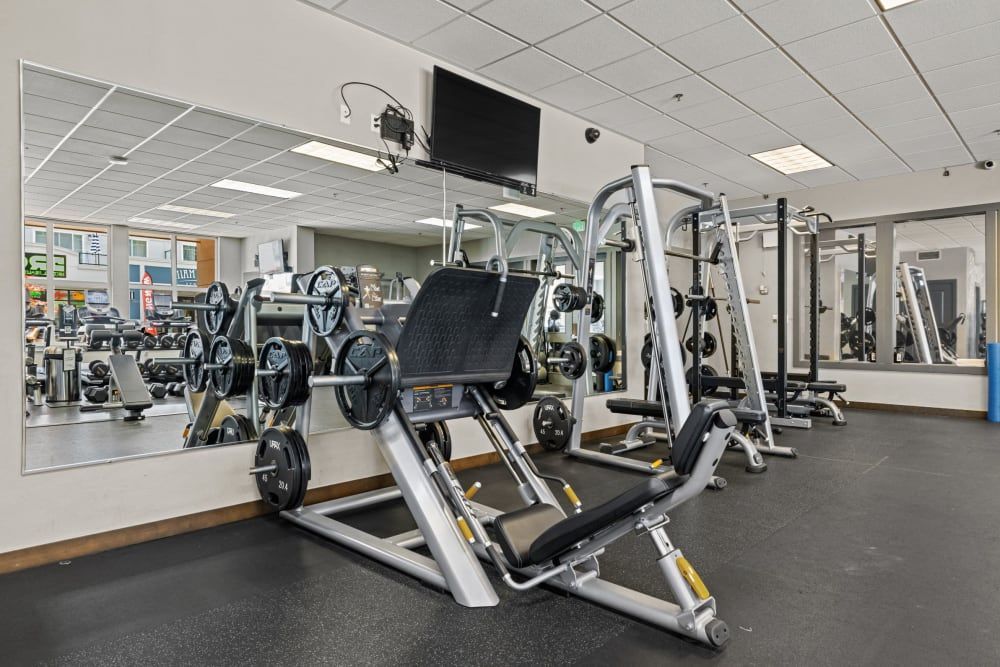 Interior of a well-equipped gym with various weight training machines including leg press and cable stations, barbell racks, and weights. The space is clean and organized, with large mirrors reflecting the equipment and a TV mounted on the wall. Outside the window, urban life is visible with cars and buildings at Copperline at Point Ruston in Tacoma, WA.