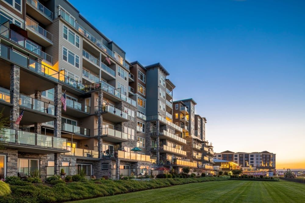 Modern multi-story apartment buildings with balconies and large windows, featuring a mix of stone and siding façade, captured at dusk with warm interior lights and a clear sky. A well-manicured lawn stretches out in the foreground at Copperline at Point Ruston in Tacoma, WA.