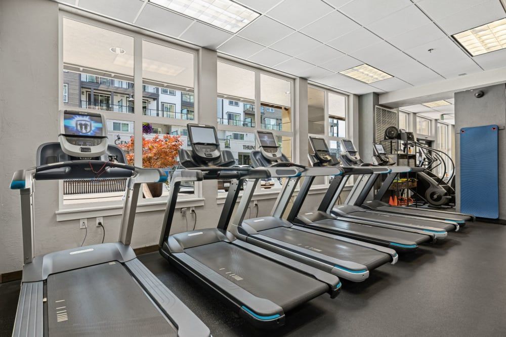 An interior view of a well-lit gym with multiple treadmills lined up in front of large windows overlooking a city street. Exercise bikes and a blue floor mat are visible in the background, to the right of the treadmills at Copperline at Point Ruston in Tacoma, WA.
