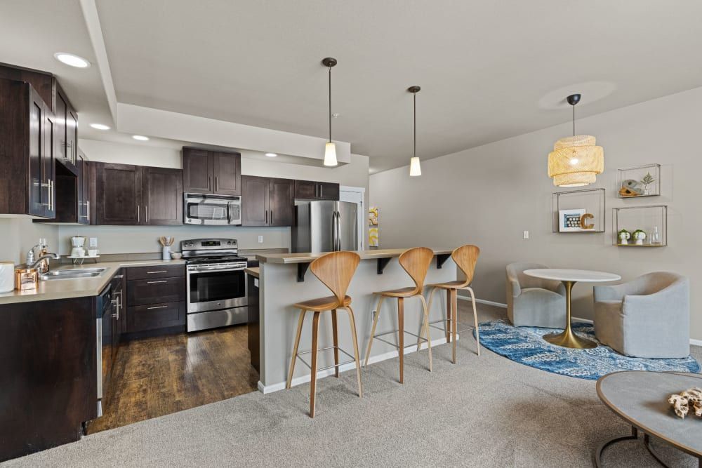 Modern kitchen with dark wood cabinets and stainless steel appliances, an island with wooden bar stools, and an adjacent dining area with a small white table, grey upholstered chairs, and decorative shelves on a wall above a blue area rug at Copperline at Point Ruston in Tacoma, WA.