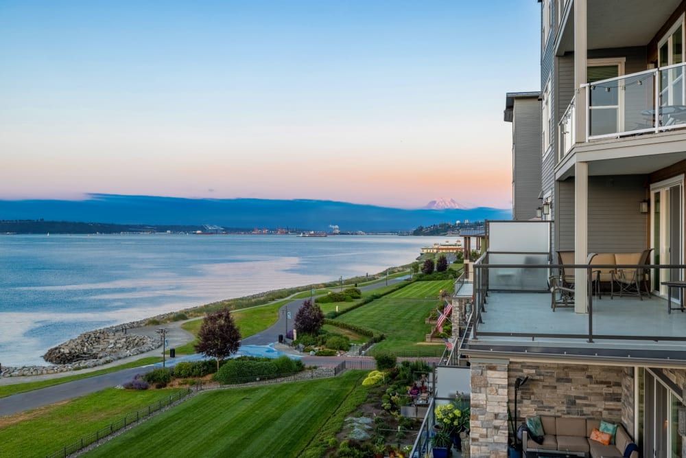 Twilight view from a residential balcony featuring well-manicured lawns leading to a serene seaside pathway beside calm waters, with a distant mountain silhouetted against the gradient sky at Copperline at Point Ruston in Tacoma, WA.