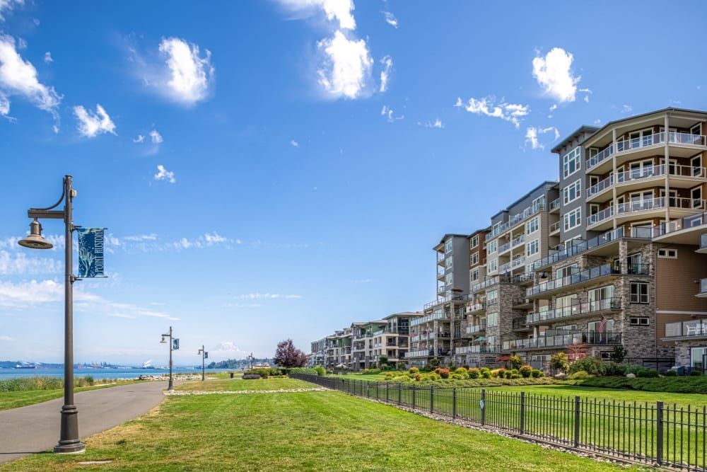 A row of modern multi-story apartment buildings with balconies overlooking a lush green lawn, bordered by a black wrought iron fence, with a paved walking path and street lamps to the left. The backdrop showcases a clear blue sky with a few clouds and distant views of a calm sea, possibly indicating a coastal location at Copperline at Point Ruston in Tacoma, WA.