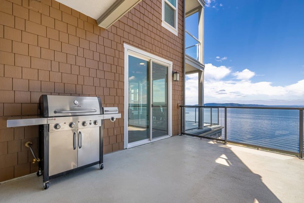A spacious balcony with a stainless steel gas grill on the left, overlooking a serene body of water under a clear blue sky. The balcony features a brown brick exterior wall to the left and glass doors that lead indoors, plus a modern glass and metal railing providing an unobstructed view of the water at Copperline at Point Ruston in Tacoma, WA.