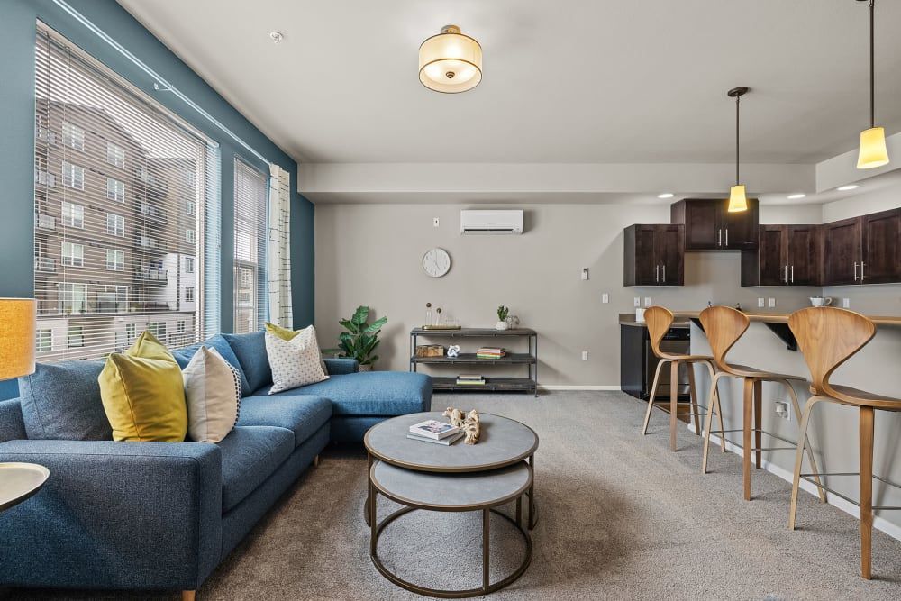 Modern living room with L-shaped blue sofa adorned with yellow and white pillows, a round coffee table, and a small side table with a plant and a lamp. To the right is a kitchen with dark wood cabinets, stainless steel appliances, and a breakfast bar with stylish wooden stools under pendant lights. Large windows offer a view of adjacent buildings, while the room features a blend of natural and artificial light at Copperline at Point Ruston in Tacoma, WA.