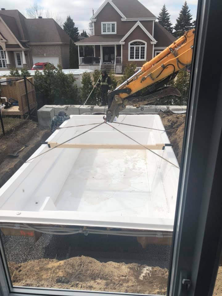 Une grande piscine blanche est soulevée par une grue devant une maison.