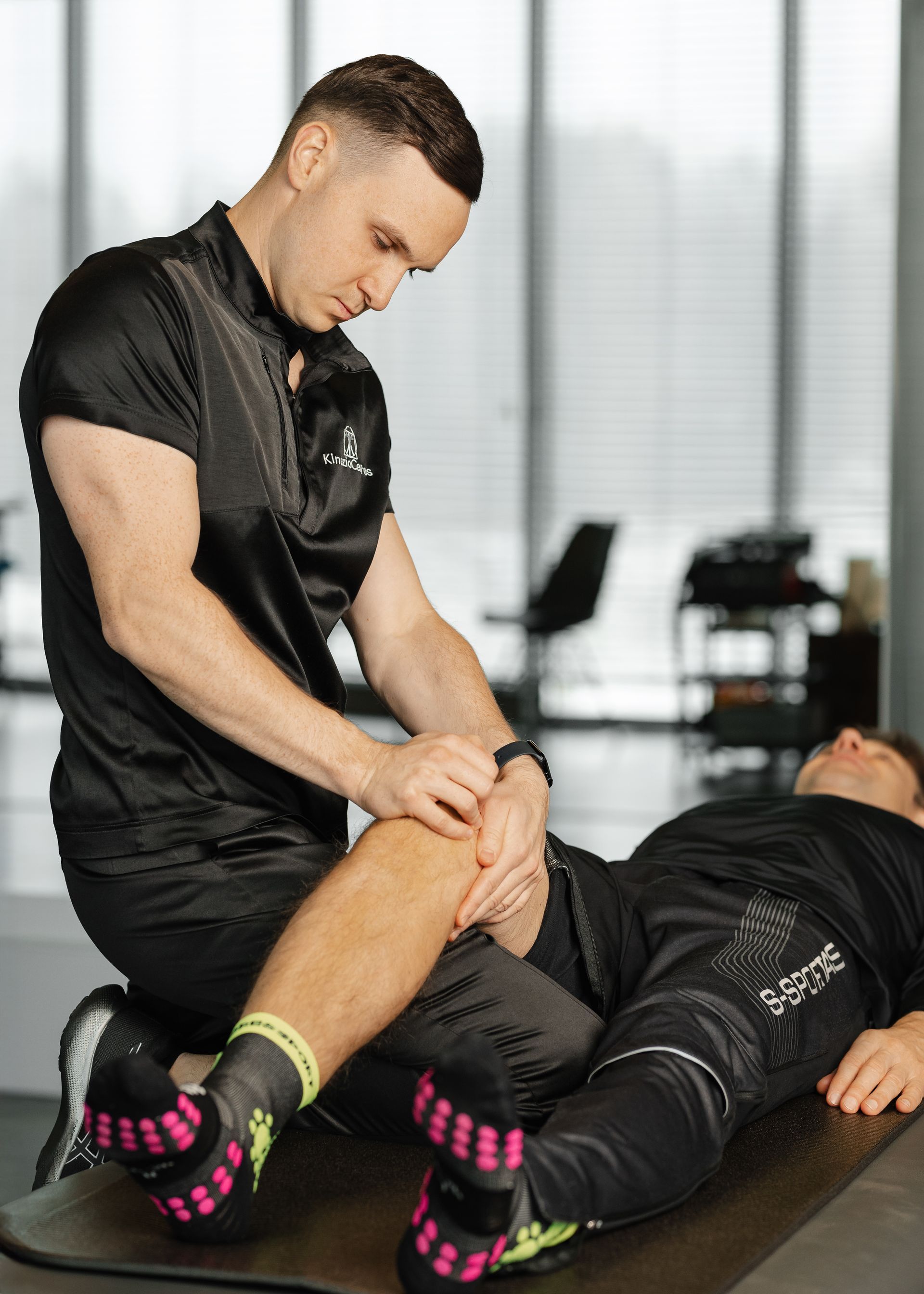 Vyras daro masažą kitam vyrui ant kilimėlio sporto salėje.