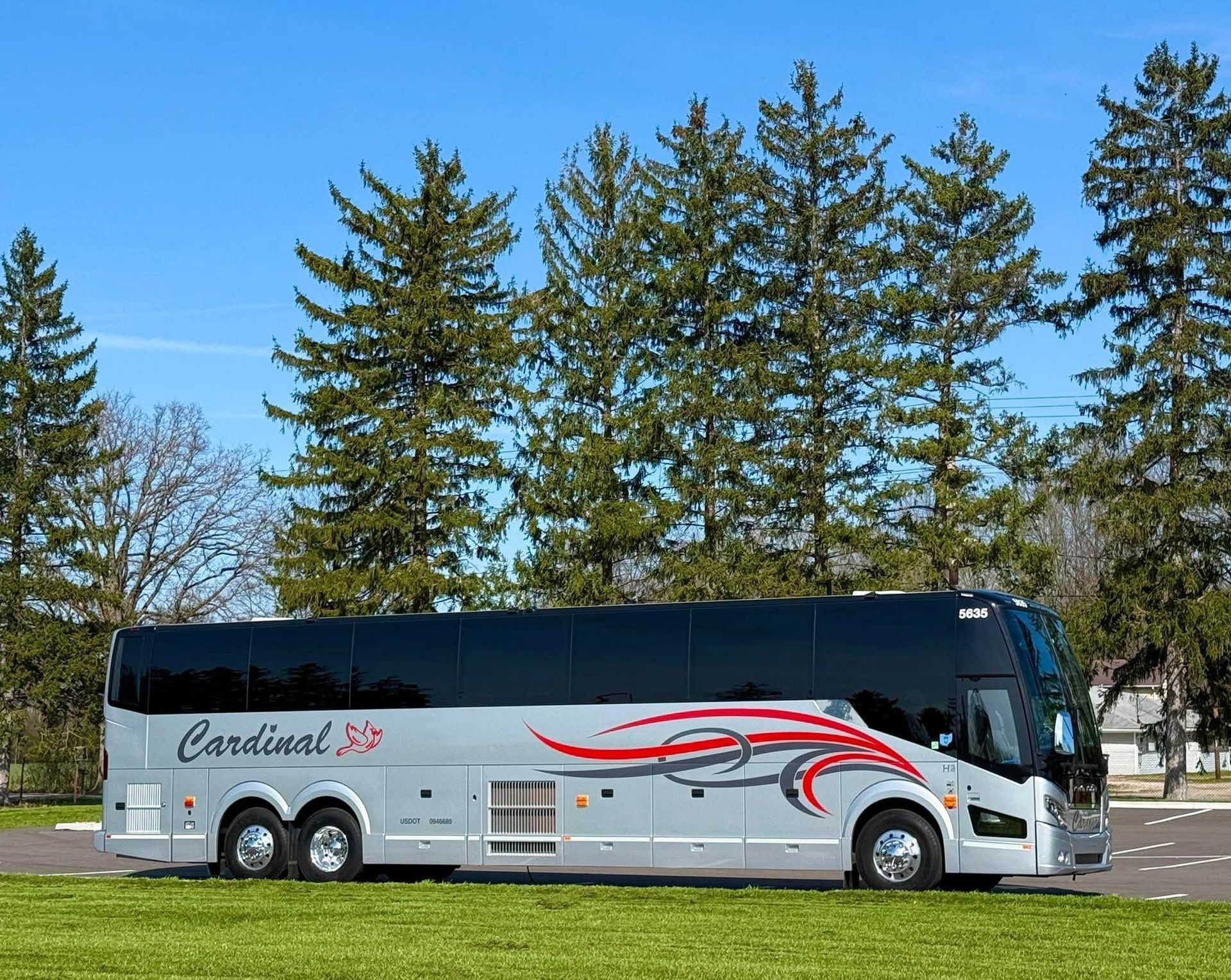 Silver charter bus parked on a grassy area with trees in the background, ready for group travel.