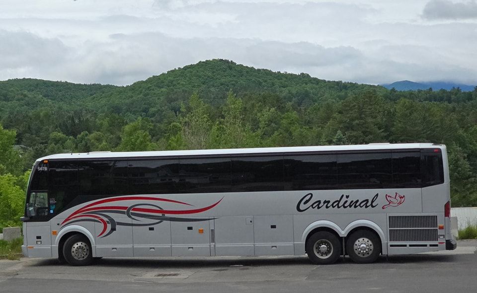 White  cardinal bus driving parked with hill and vegetation in the background.