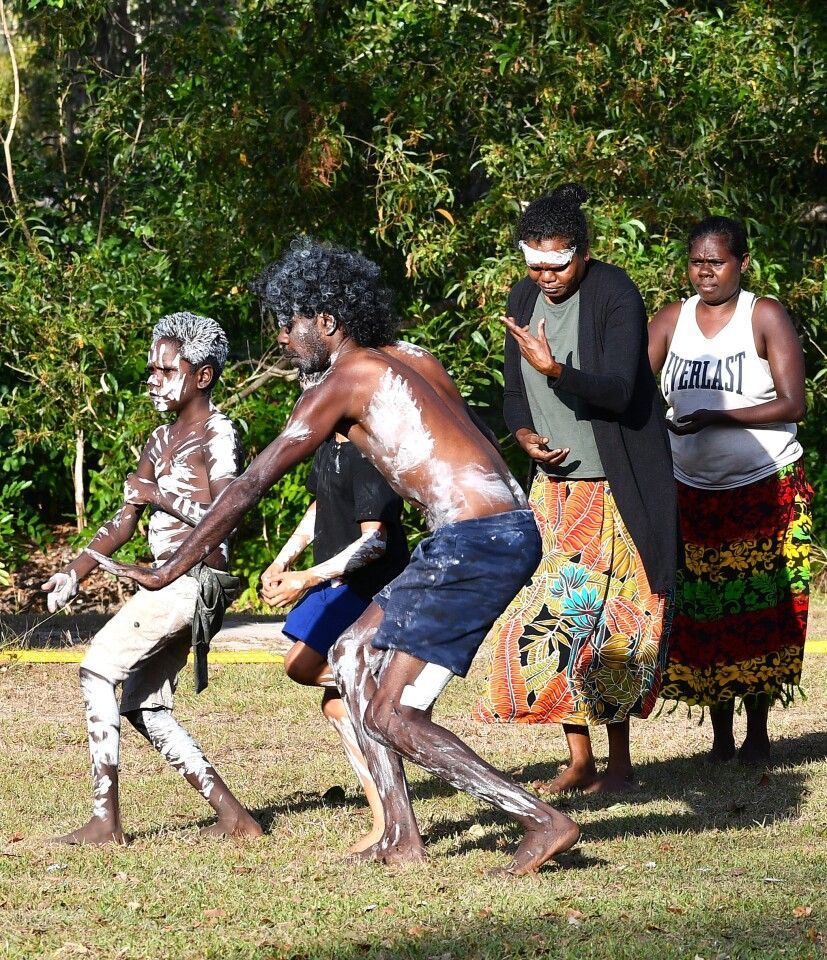 Indigenous dancers in white paint perform on grass, observed by two others.