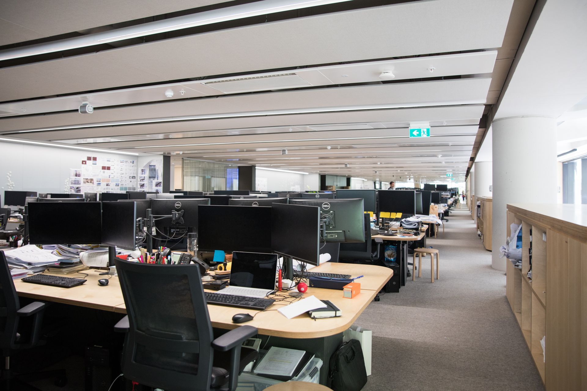 Office workspace with rows of desks, computers, chairs, and overhead lighting.