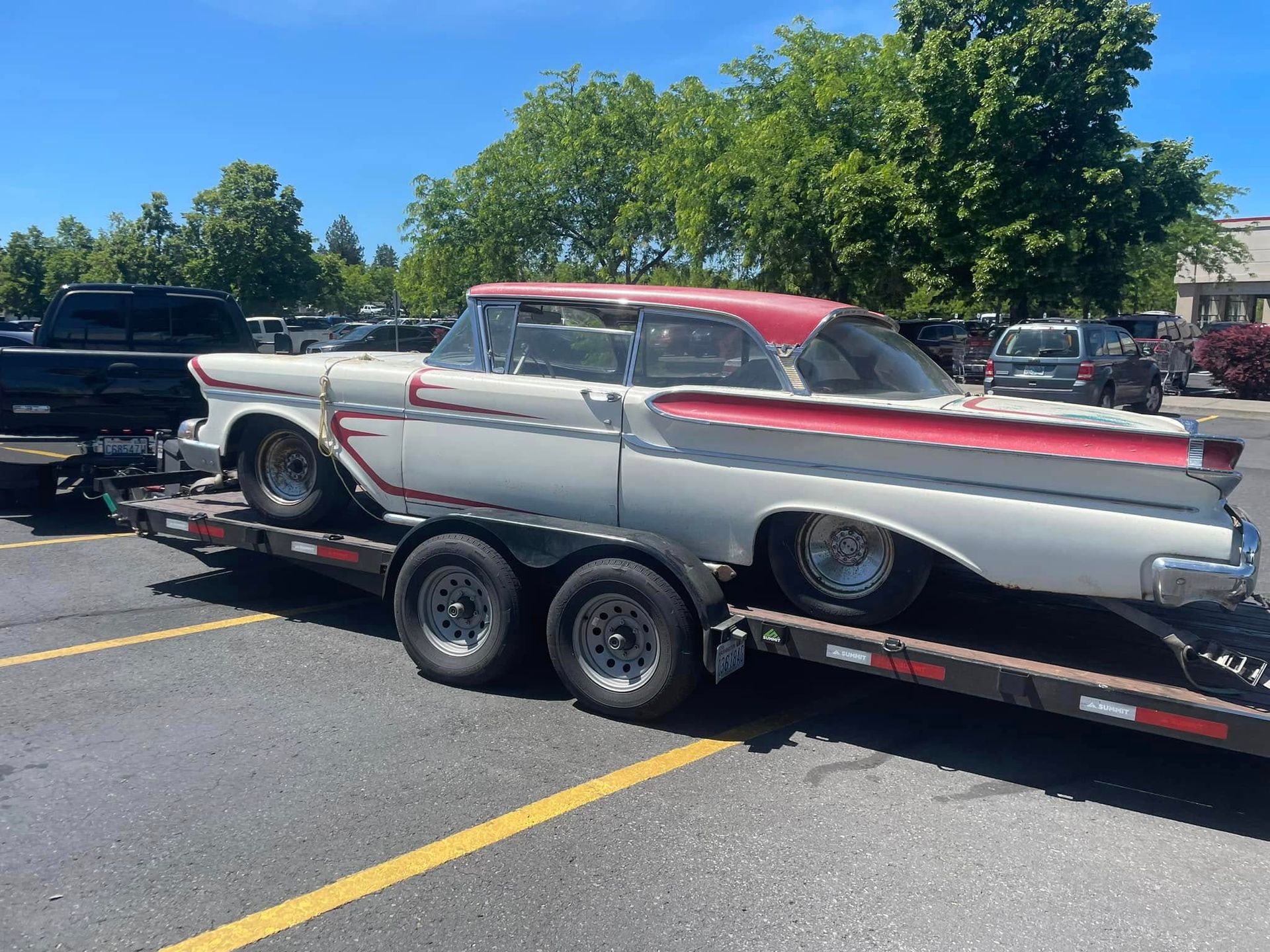 A white and red classic car loaded onto a flatbed trailer in a parking lot under a bright blue sky.