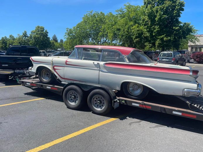 A white and red classic car loaded onto a flatbed trailer in a parking lot under a bright blue sky.