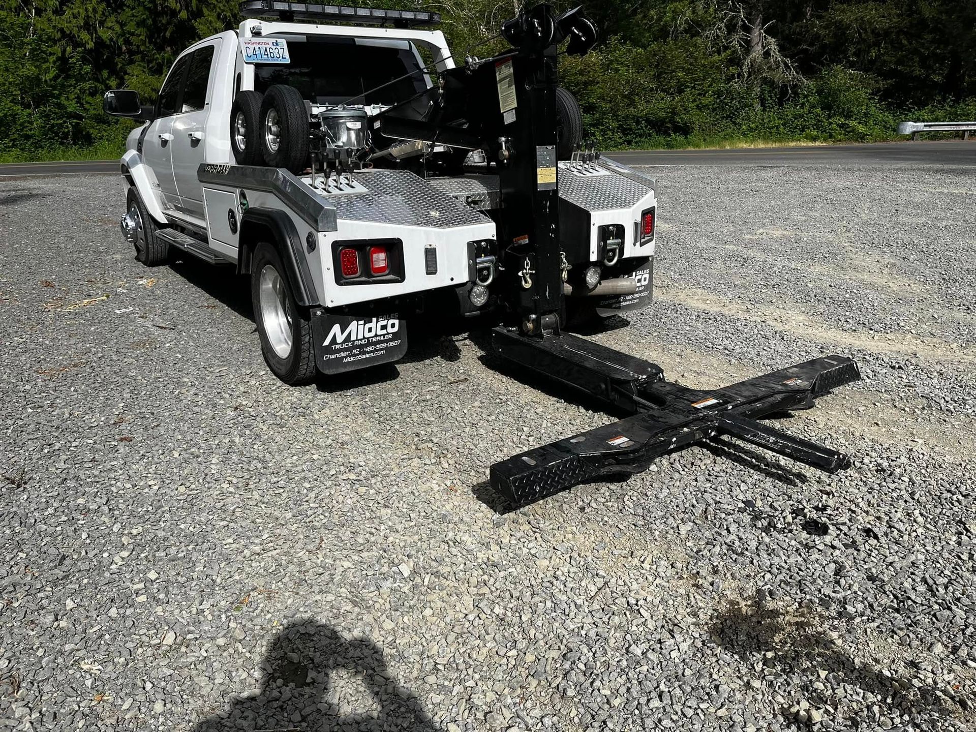A white tow truck parked on a gravel lot with its extended metal towing boom lowered toward the ground.