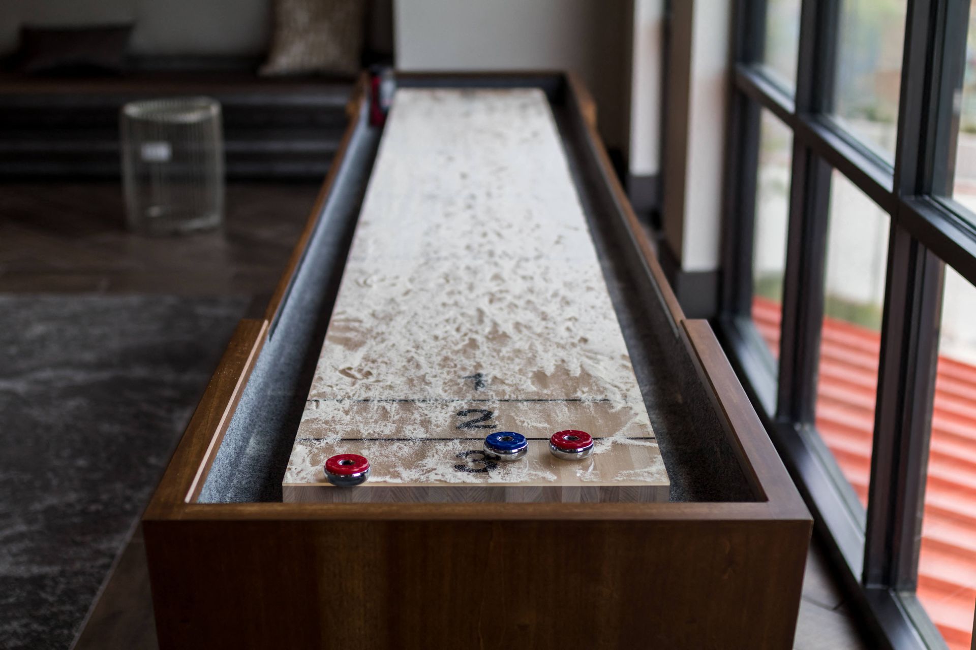 A shuffleboard table is sitting in front of a window in the game room.