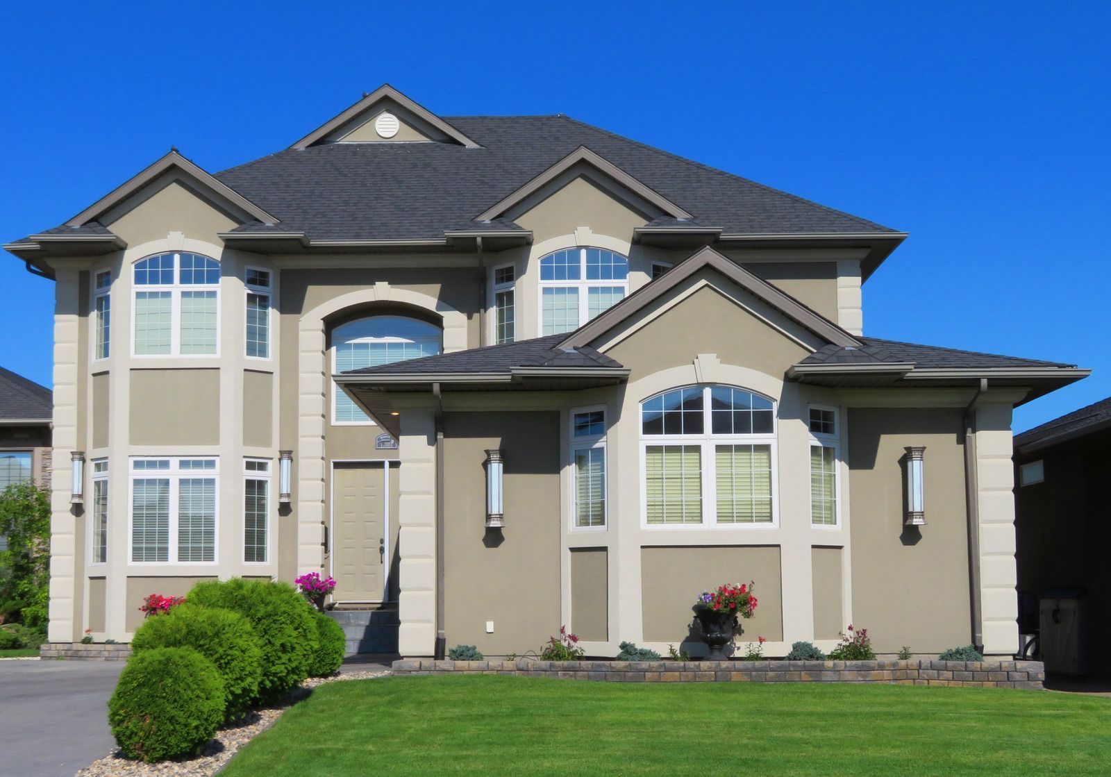 Two-story beige house with dark gray roof, arched windows, and green lawn under a clear blue sky.