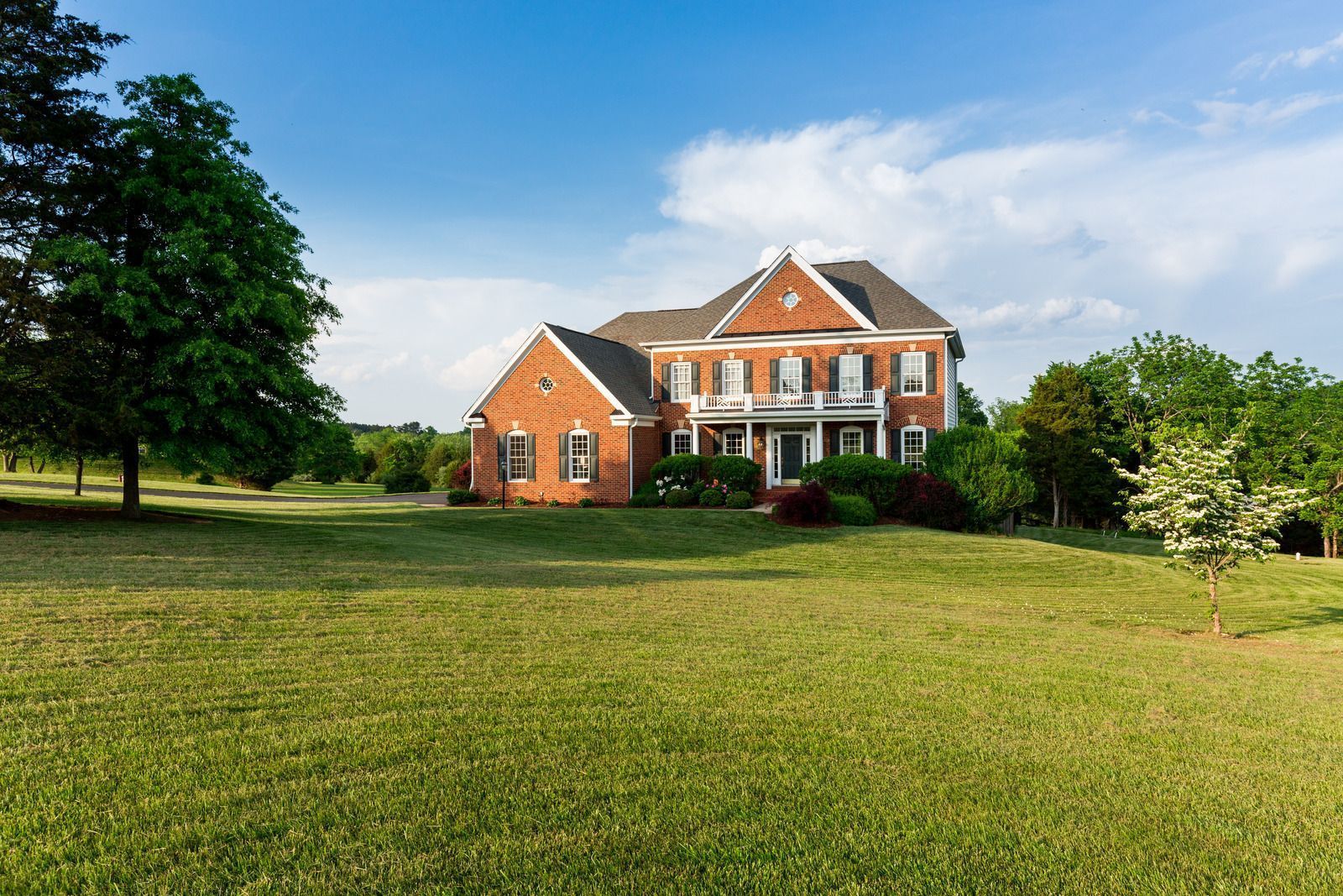 Brick house with white shutters and columns on a large, grassy lawn under a blue sky.