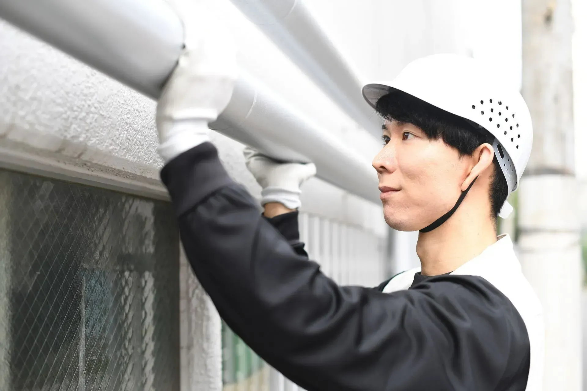 Man in hard hat and gloves installing a gutter on a building's exterior.