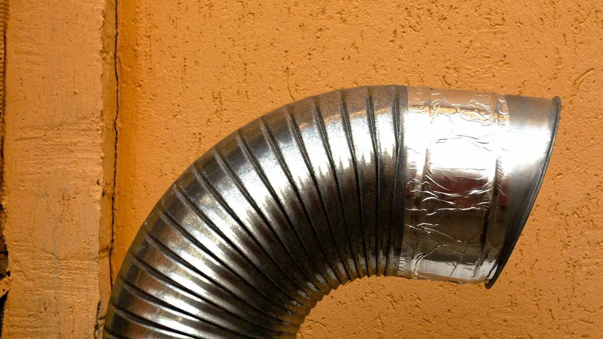 Curved, silver, flexible ductwork against a textured orange wall.