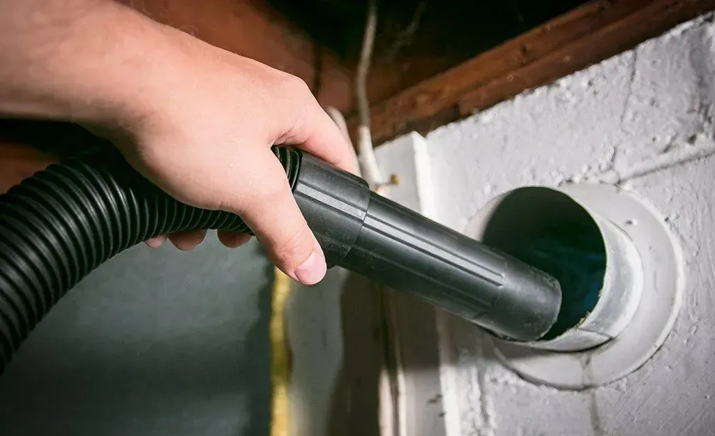 Person using a vacuum hose to clean a white dryer vent in a basement.