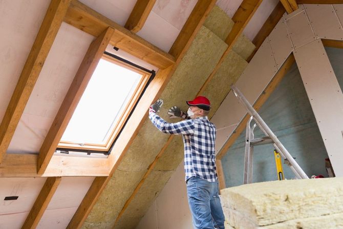Worker in a red hat installing insulation in an attic with exposed wooden beams and a skylight